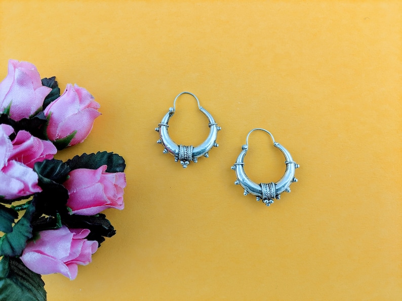 May include: A pair of silver hoop earrings with a decorative design. The earrings are on a yellow background.