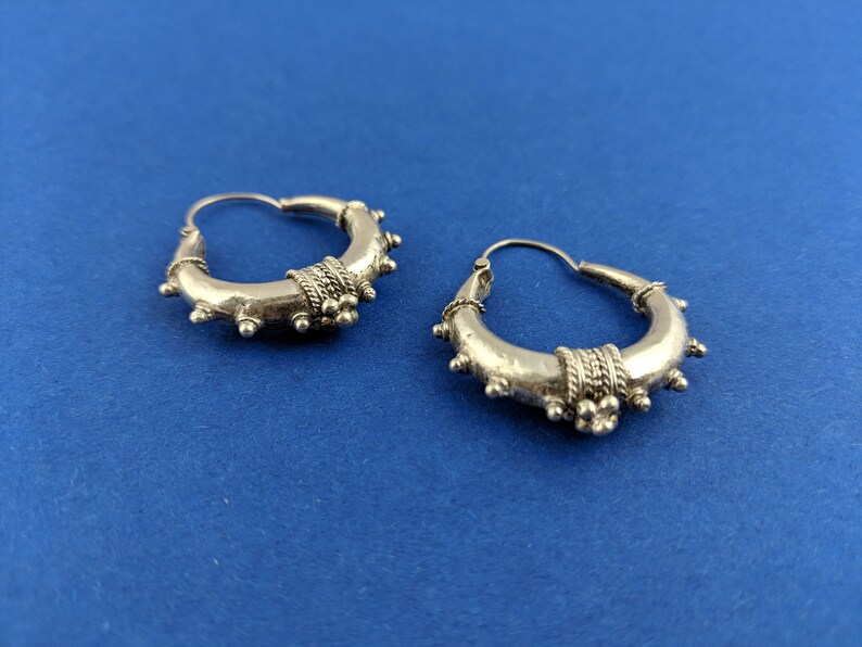May include: A pair of silver hoop earrings with a textured design. The earrings have a circular shape and are decorated with small, raised dots and a band of intricate detail.