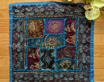 Puede incluir: Una almohada cuadrada de patchwork azul y morado con bordados intrincados y lentejuelas. La almohada tiene un borde azul y está hecha de una variedad de telas.