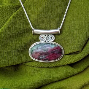 May include: A silver chain necklace with a pendant featuring a green and pink oval gemstone set in a silver frame. The pendant has a decorative silver bail with a swirl design.