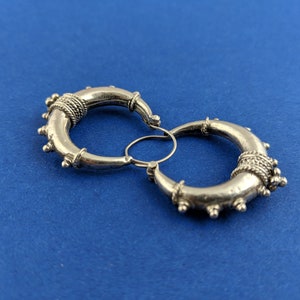 May include: A pair of silver hoop earrings with intricate detailing. The hoops are thick and have a textured surface. The earrings are connected by a small silver loop.