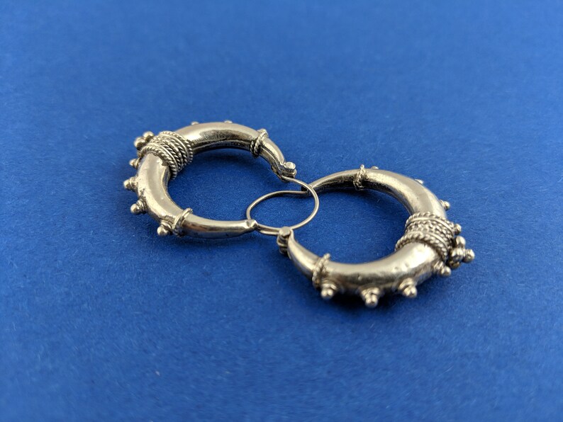 May include: A pair of silver hoop earrings with intricate detailing. The hoops are thick and have a textured surface. The earrings are connected by a small silver loop.