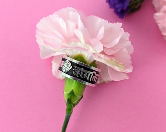Puede incluir: Un anillo de plata con una banda negra y un diseño floral. La banda está inscrita con el texto "Om Mani Padme Hum". El anillo está apoyado en un clavel rosa.