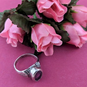 May include: A silver ring with a purple gemstone set in a decorative band. The ring is lying on a pink surface with a bouquet of pink silk roses.