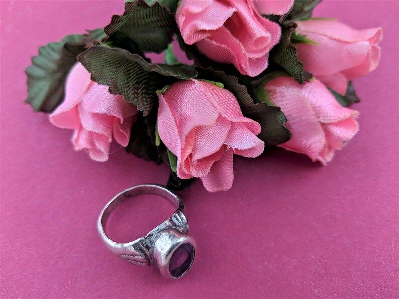 May include: A silver ring with a purple gemstone set in a decorative band. The ring is lying on a pink surface with a bouquet of pink silk roses.