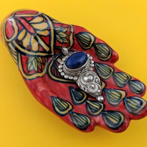 May include: A ceramic hand painted in red, yellow, and blue with a detailed floral pattern. The hand is holding a silver pendant with a blue gemstone.