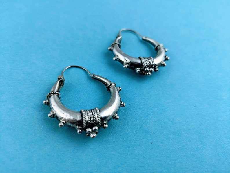 May include: A pair of silver hoop earrings with a decorative design. The earrings have a circular shape and are adorned with small beads and a textured band.