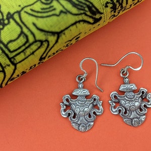 May include: A pair of silver earrings with intricate designs. Each earring features a stylized, ornate motif with a central circular element and flowing, curved lines. The earrings are suspended from simple silver hooks.