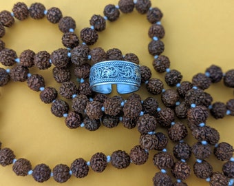 Puede incluir: Un anillo de plata con un intrincado diseño floral, descansando sobre un collar de cuentas marrones.