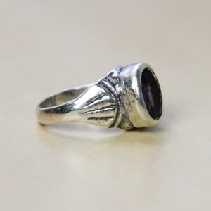May include: A silver ring with a purple gemstone set in a bezel setting. The ring has a decorative band with a textured design.