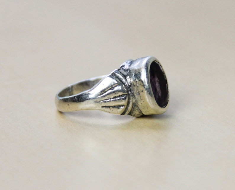 May include: A silver ring with a purple gemstone set in a bezel setting. The ring has a decorative band with a textured design.
