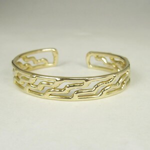 May include: A gold-colored cuff bracelet with an open design. The bracelet features a repeating wave-like pattern across its surface. The open design allows for easy wear and removal. This piece of jewelry is suitable for everyday wear or special occasions.