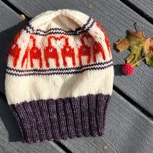 May include: A knitted winter hat with a cream-colored base, featuring a pattern of red figures. The hat has a purple ribbed brim and is decorated with a leaf and a red berry.