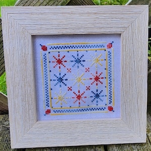 May include: A white wooden frame with a white mat and a cross-stitch design of red, blue, and yellow stars on a white background.