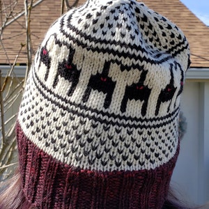 May include: A white and black knit hat with a burgundy brim. The hat features a repeating pattern of black cats with red eyes.