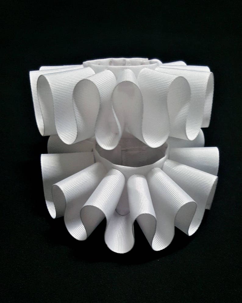 May include: A white neck ruff featuring two stacked tiers of pleated grosgrain ribbon. The accessory is constructed with a circular base and structured, undulating folds that create a voluminous, ruffled appearance around the central opening.