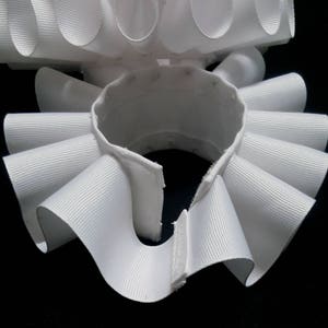 May include: A white, circular, pleated neck ruff made of ribbed fabric. The accessory features a series of uniform, folded loops arranged in a continuous ring, with a small opening at the front secured by a hook and eye closure mechanism.