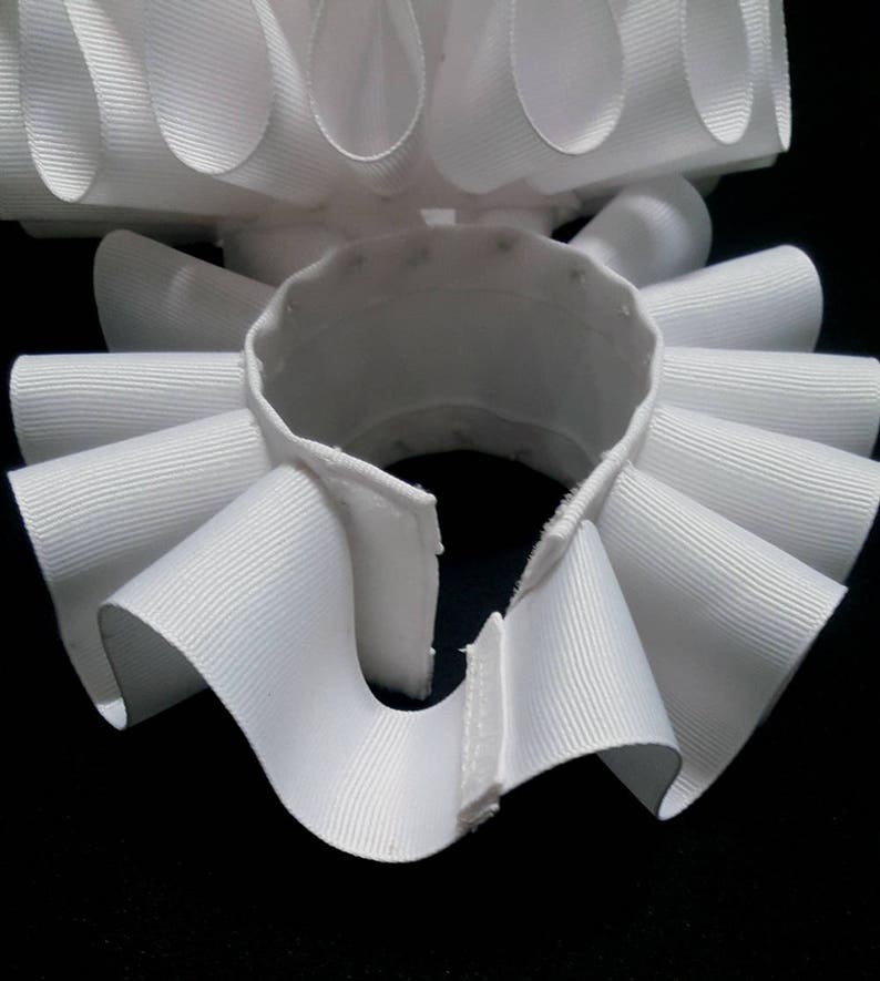 May include: A white, circular, pleated neck ruff made of ribbed fabric. The accessory features a series of uniform, folded loops arranged in a continuous ring, with a small opening at the front secured by a hook and eye closure mechanism.
