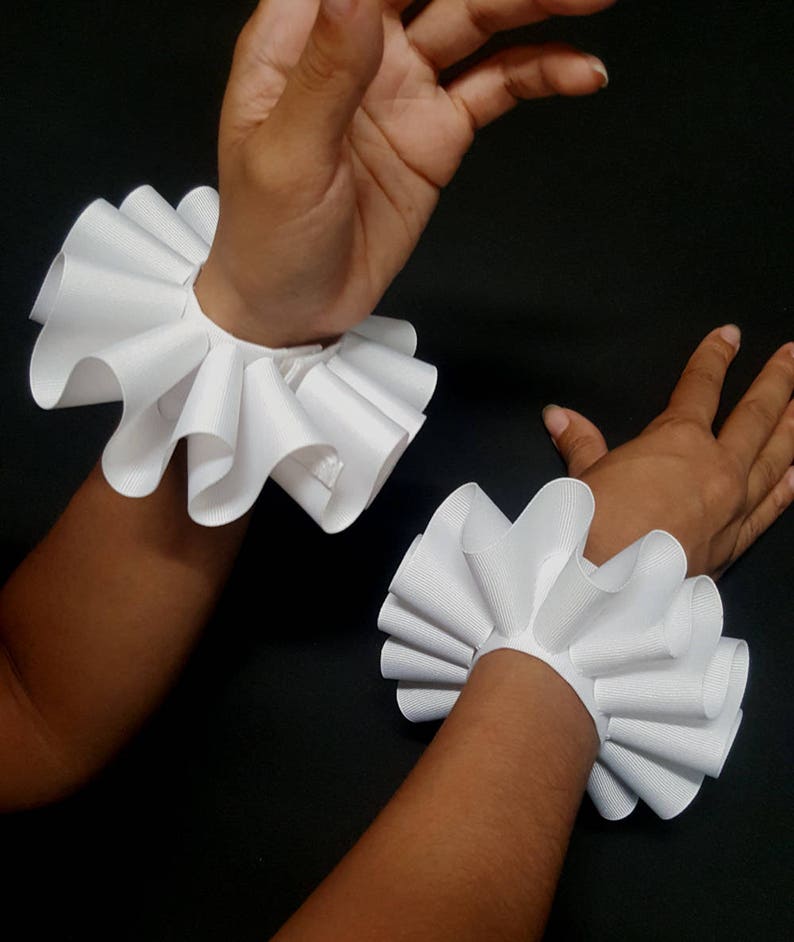 May include: A pair of white wrist ruffles made from a ribbed fabric. Each accessory features a circular band with a wide, pleated, and flared edge that creates a voluminous, wavy appearance around the wrist area, providing a decorative accent.