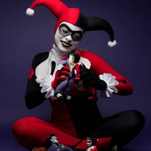 May include: A costume featuring a red and black jester suit with a matching two-toned hood, white ruffled collar and cuffs, and white face makeup with a black eye mask. Two small dolls are held in the hands, and two more sit on the floor.