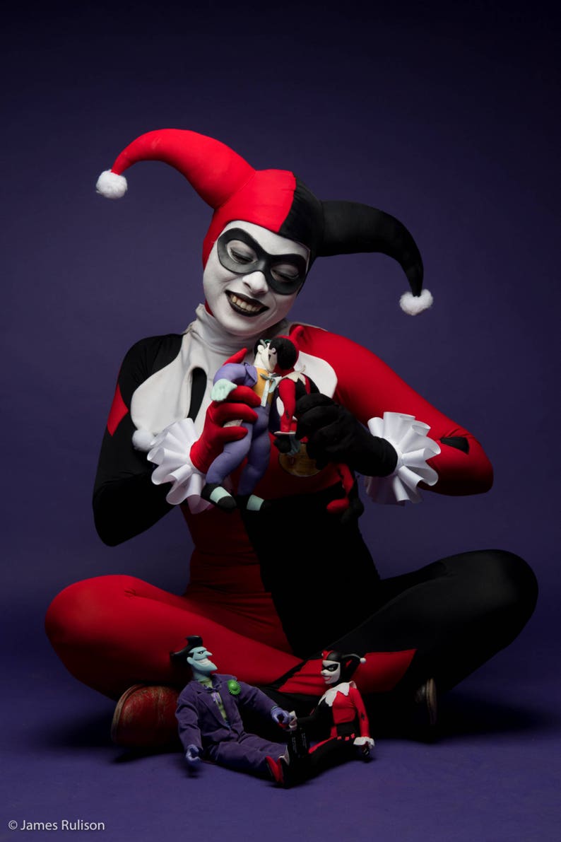 May include: A costume featuring a red and black jester suit with a matching two-toned hood, white ruffled collar and cuffs, and white face makeup with a black eye mask. Two small dolls are held in the hands, and two more sit on the floor.