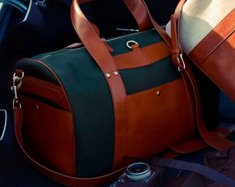 Puede incluir: Una bolsa de viaje de cuero marrón y verde oscuro con asa superior y bandolera. La bolsa está en la parte trasera de un coche.