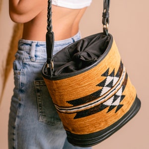 Può includere: Borsa a mano cilindrica con esterno tessuto e strutturato nei toni dell'oro, nero e bianco. La borsa presenta un motivo geometrico e una base e finiture in pelle nera. Ha un interno nero e una tracolla in pelle nera con dettagli dorati.