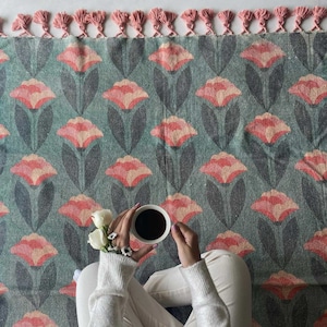Può includere: Un tappeto color turchese con un motivo floreale con fiori rosa e foglie verde scuro. Una persona è seduta sul tappeto con una tazza di caffè in mano.