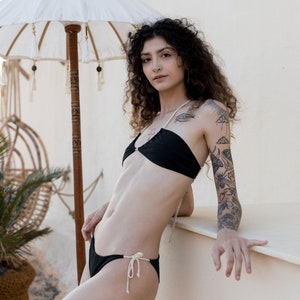 Peut inclure: Une femme avec des tatouages sur le bras porte un haut de bikini noir et un bas de bikini assorti avec des liens blancs. Elle se tient devant un mur blanc avec un parasol blanc derrière elle.