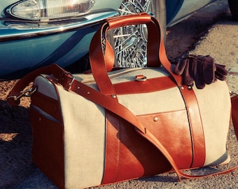 Puede incluir: Una bolsa de viaje de cuero marrón y beige con una correa larga. La bolsa está sentada sobre una superficie gris junto a un coche clásico.