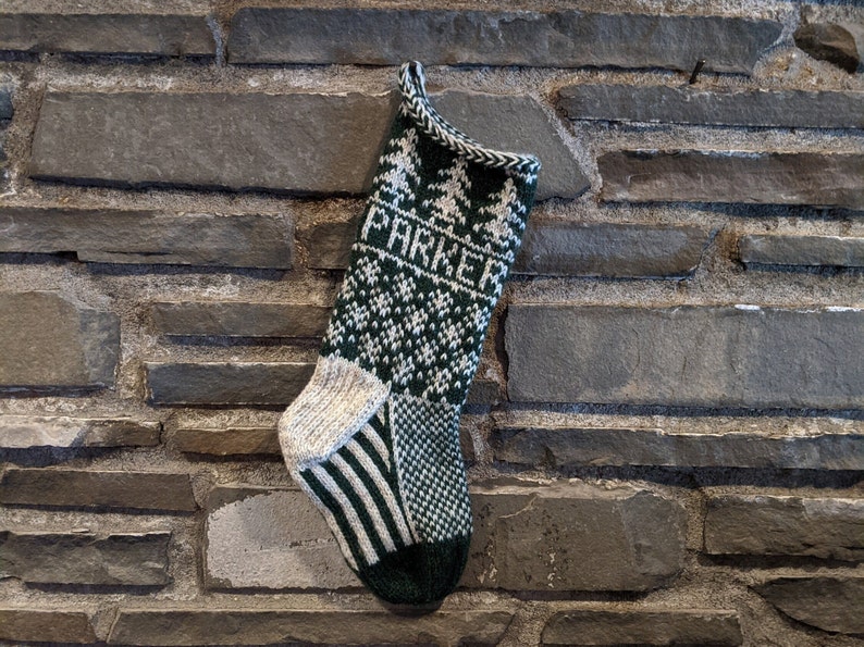 Christmas stocking knit with green and white yarn hangs on a grey stone wall