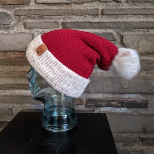 May include: A red and white knit hat with a white faux fur pom-pom. The hat is displayed on a glass head. The hat has a brown leather tag attached to the brim. The hat is a festive winter accessory.