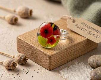 May include: A ring featuring a round, clear fused glass cabochon with embedded red flower petals and green leaves. The ring has a silver-toned metal band and is accompanied by a brown paper tag with the text HANDMADE FUSED GLASS printed on it.