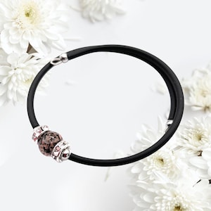 Puede incluir: Una pulsera de silicona negra con un dije de plata que presenta una piedra moteada de color rosa y marrón. El dije está acentuado con diamantes de imitación transparentes.