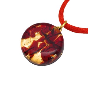 Peut inclure: Un pendentif rond avec un motif rouge et or, suspendu à un cordon rouge. Le pendentif a une bélière dorée. Le rouge est translucide, révélant un motif de feuille d'or à l'intérieur du verre. Le cordon est fin et lisse.