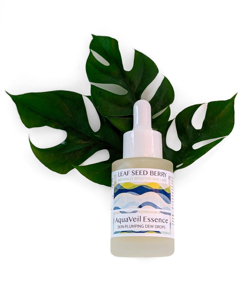 May include: A clear glass bottle of AquaVeil Essence skin-plumping dew drops with a white dropper top. The bottle is labeled with the text "LEAF SEED BERRY" and "NATURALLY EFFECTIVE SKIN CARE". The bottle is set against a backdrop of large green leaves.