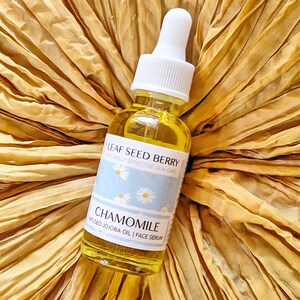 May include: A glass bottle of chamomile infused jojoba oil face serum with a white dropper top. The bottle has a blue and white label with the text "Leaf Seed Berry Naturally Effective Skin Care Chamomile Infused Jojoba Oil | Face Serum".