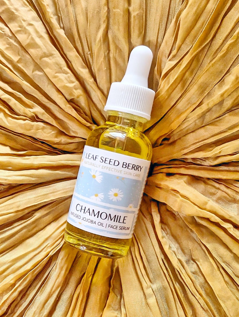 May include: A glass bottle of chamomile infused jojoba oil face serum with a white dropper top. The bottle has a blue and white label with the text "Leaf Seed Berry Naturally Effective Skin Care Chamomile Infused Jojoba Oil | Face Serum".