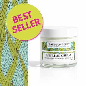 Peut inclure: Un pot en verre transparent de crème hydratante Leaf Seed Berry Mermaid Cream Hyaluronic Marine avec un couvercle blanc. L'étiquette présente un motif de feuilles vertes et bleues et indique 2 fl oz. Un cercle jaune avec du texte rose indique BEST SELLER.