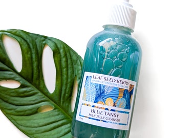 May include: A teal-colored bottle of Blue Tansy Milk Jelly Cleanser with a white pump dispenser. The label reads "Leaf Seed Berry, Naturally Effective Skin Care." A large green leaf is next to the bottle. The bottle contains 4 fl oz.