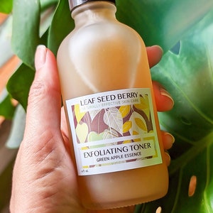 Pu&ograve; includere: Una bottiglia di vetro trasparente con un tappo marrone che contiene un tonico esfoliante all'essenza di mela verde. La bottiglia ha un'etichetta con il testo "LEAF SEED BERRY NATURALLY EFFECTIVE SKIN CARE EXFOLIATING TONER GREEN APPLE ESSENCE 4 fl. oz."
