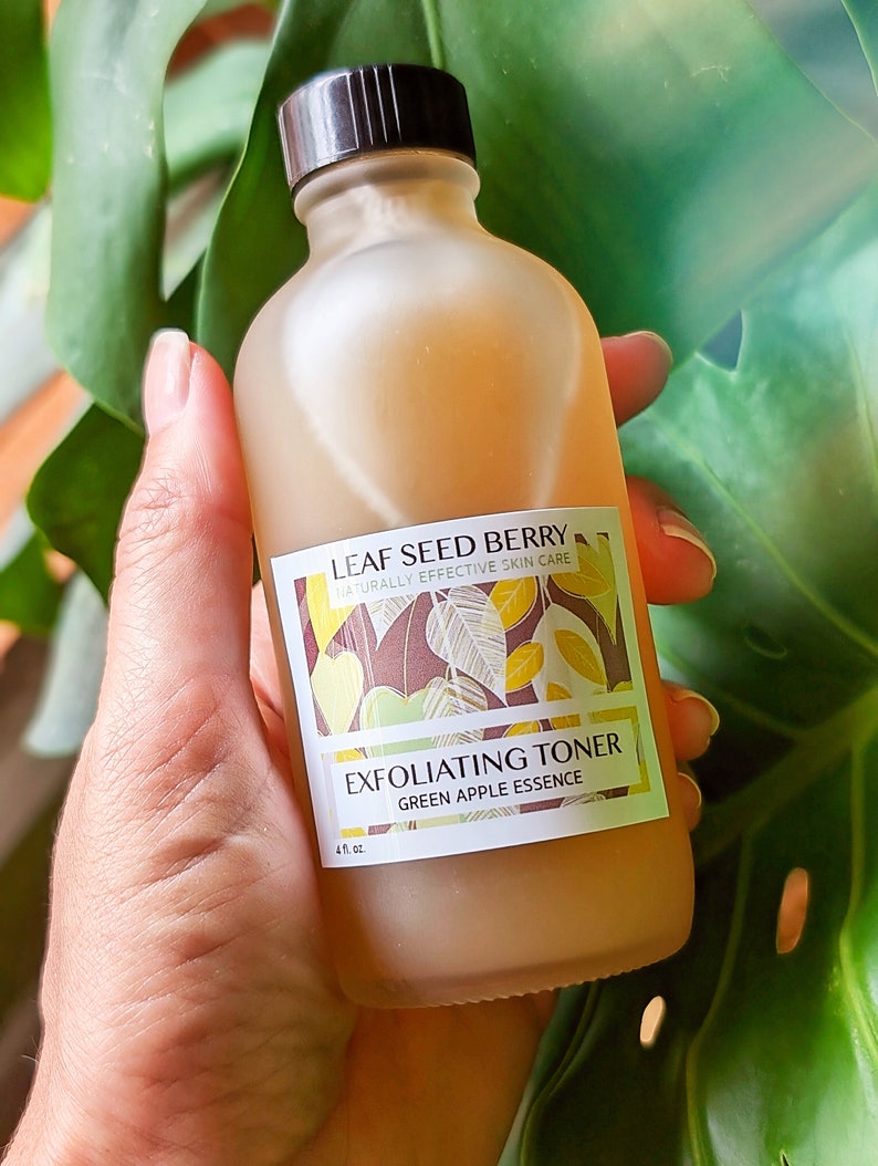 Pu&ograve; includere: Una bottiglia di vetro trasparente con un tappo marrone che contiene un tonico esfoliante all'essenza di mela verde. La bottiglia ha un'etichetta con il testo "LEAF SEED BERRY NATURALLY EFFECTIVE SKIN CARE EXFOLIATING TONER GREEN APPLE ESSENCE 4 fl. oz."