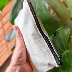 May include: A plain white canvas zipper pouch with a black zipper. The pouch is held in a hand.