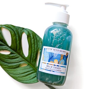 May include: A blue and white bottle of Leaf Seed Berry Blue Tansy Milk Jelly Cleanser. The bottle has a pump dispenser and a label with a tropical leaf design.