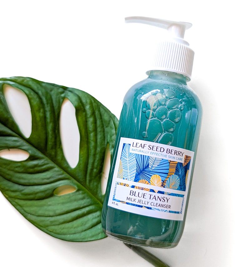 May include: A blue and white bottle of Leaf Seed Berry Blue Tansy Milk Jelly Cleanser. The bottle has a pump dispenser and a label with a tropical leaf design.