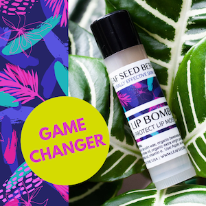 May include: A white tube of lip balm with a black cap and a blue and pink label that says "Leaf Seed Berry Naturally Effective Skin Lip Bomb Protect Lip Moisture". The label also includes ingredients and a website address. The tube is sitting on a green leafy plant.