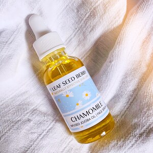 May include: A clear glass bottle with a dropper top filled with a yellow liquid. The bottle has a white label with the text "LEAF SEED BERRY NATURALLY EFFECTIVE SKIN CARE CHAMOMILE INFUSED JOJOBA OIL | FACE SERUM".