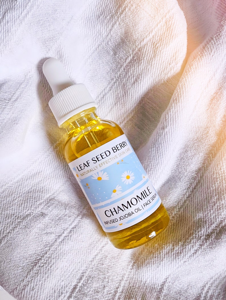 May include: A clear glass bottle with a dropper top filled with a yellow liquid. The bottle has a white label with the text "LEAF SEED BERRY NATURALLY EFFECTIVE SKIN CARE CHAMOMILE INFUSED JOJOBA OIL | FACE SERUM".