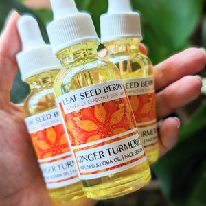 May include: Three small glass bottles of face serum are held in a hand. The bottles have white droppers and labels with orange floral designs and the text "Ginger Turmeric Infused Jojoba Oil | Face Serum". The serum is yellow.