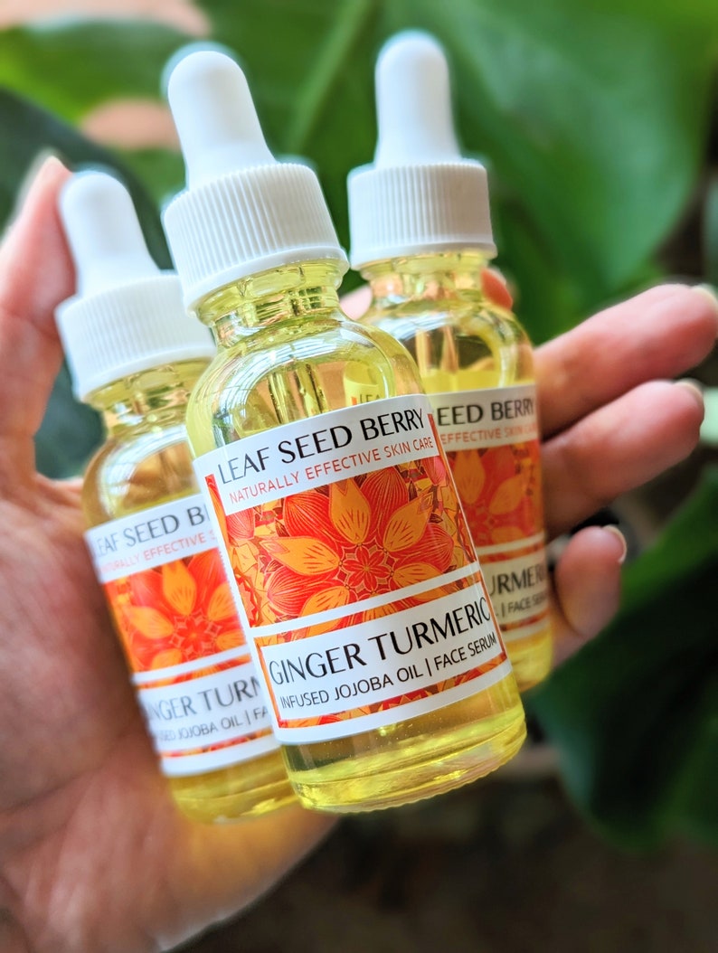 May include: Three small glass bottles of face serum are held in a hand. The bottles have white droppers and labels with orange floral designs and the text "Ginger Turmeric Infused Jojoba Oil | Face Serum". The serum is yellow.
